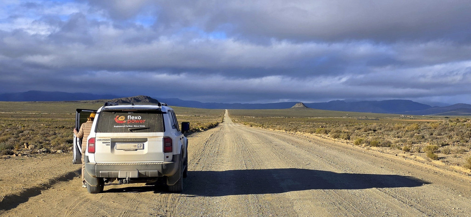 A white off-road SUV parked on a dirt road in a vast desert landscape under a dramatic cloudy sky. The rear of the vehicle is visible, with a person standing by the open driver-side door.