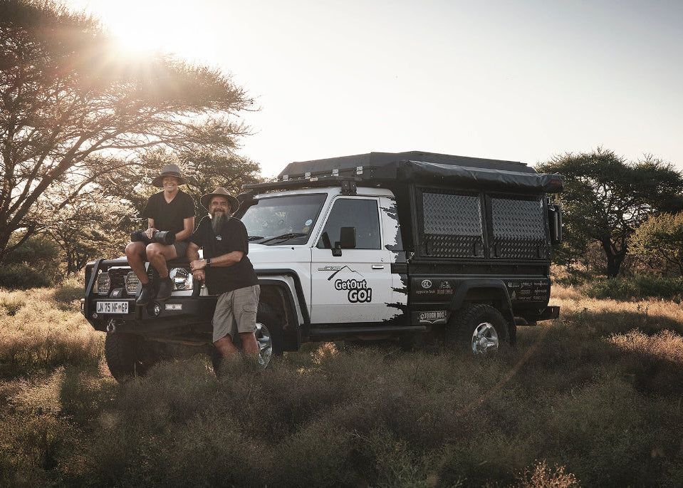 Two people standing next to a white and black off-road vehicle in a natural setting with trees and sunlight.